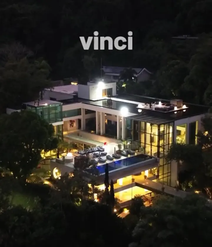 Vista aérea noturna de uma casa moderna na mata, com iluminação externa, piscina azul e jardim acesos.