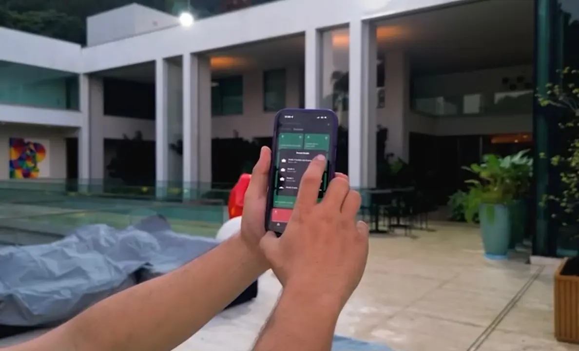 Mãos usando o app de cenas no celular, no pátio da piscina de uma casa com colunas brancas.