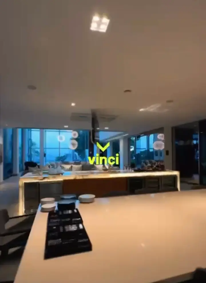 Cozinha integrada à sala, com bancada de ônix iluminada, TV suspensa no forro e janelas de vidro ao entardecer.