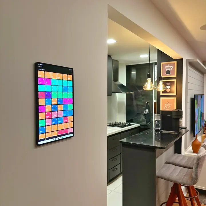 Tablet fixado na parede com dezenas de cenas coloridas, ao lado de uma cozinha com bancada de granito, pendentes e banquetas.