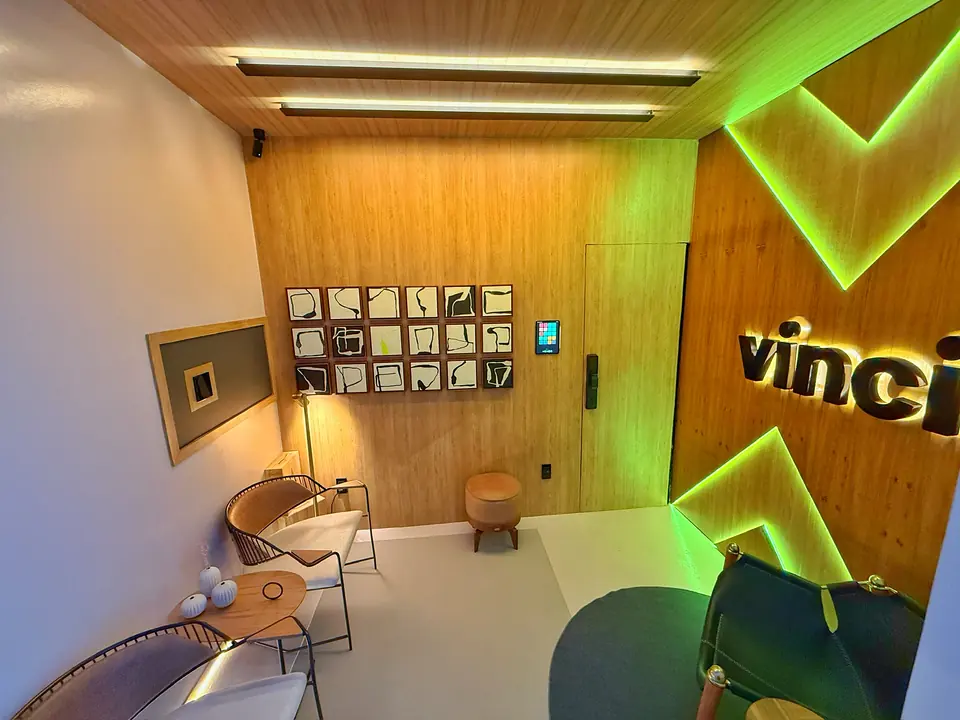 Espaço Vinci com paredes de madeira, perfis de LED no teto, tablet de controle, fechadura digital e o logo vinci com chevrons iluminados em verde.