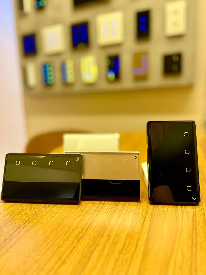 Três interruptores touch de vidro sobre uma mesa de madeira, com um mostruário de teclados ao fundo.