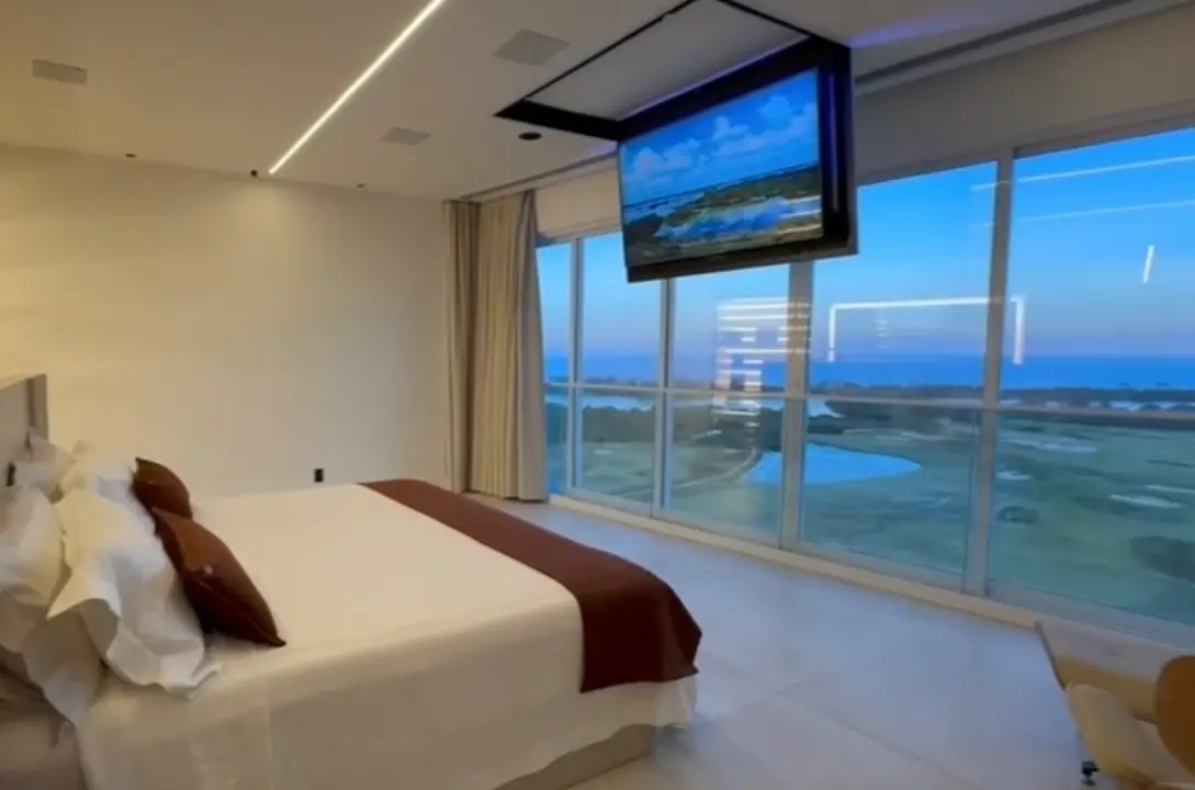 Quarto com cama de casal, TV suspensa que desce do forro e janelas do chão ao teto com vista para o mar ao entardecer.