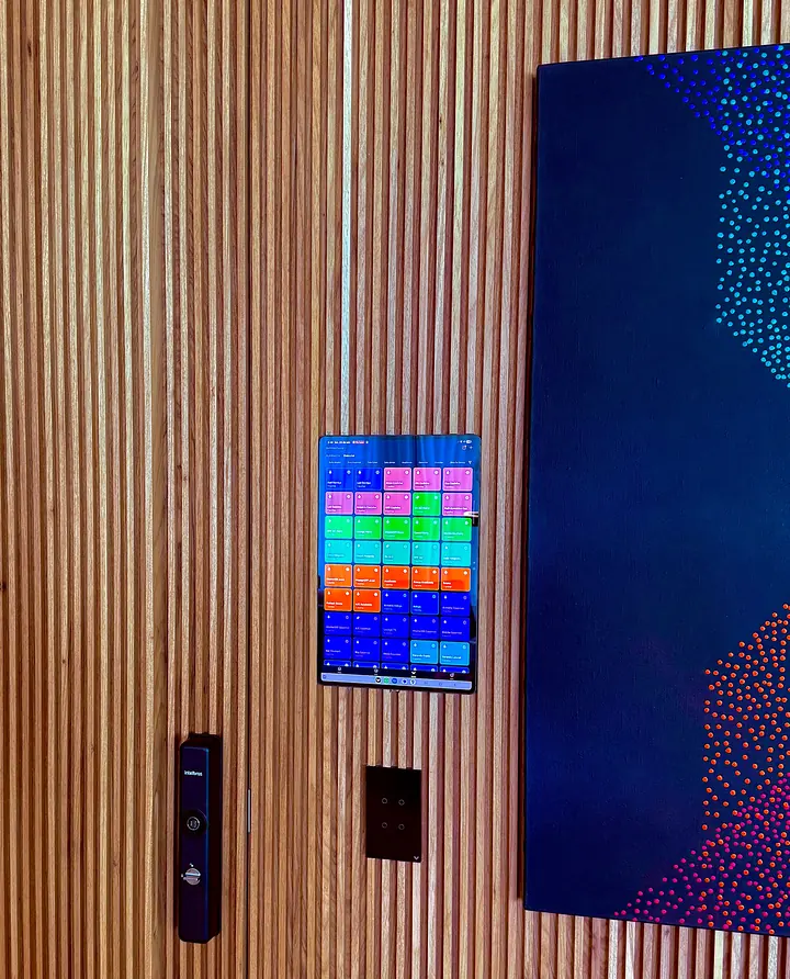 Parede de ripado de madeira com tablet de controle, interruptor touch preto e fechadura digital, ao lado de um quadro azul.