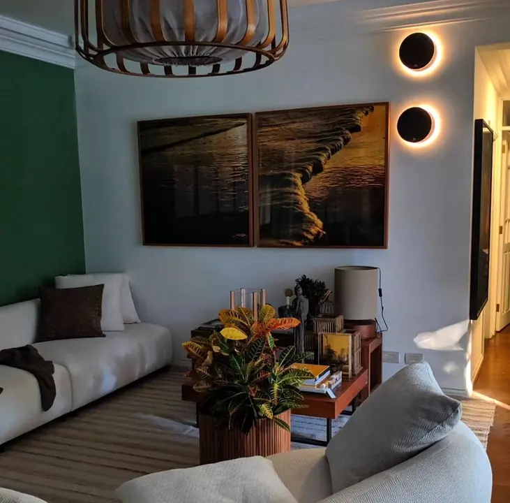 Sala de estar com parede verde, sofá claro, dois quadros e arandelas redondas de luz indireta.