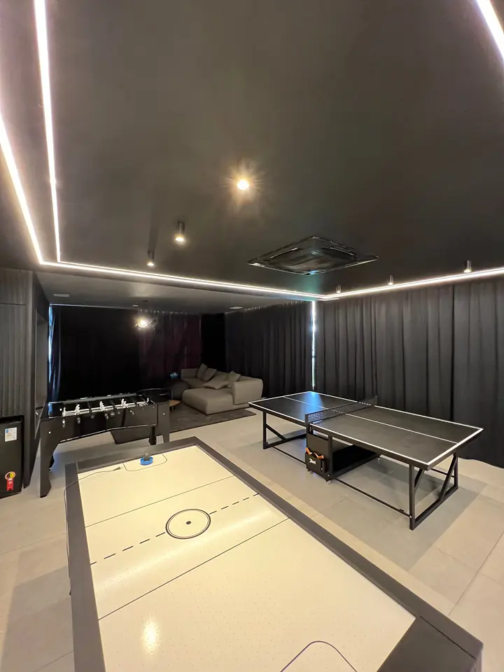 Sala de jogos escura com perfis de LED no forro, mesas de pingue-pongue, aero-hockey e pebolim e cortinas pretas.