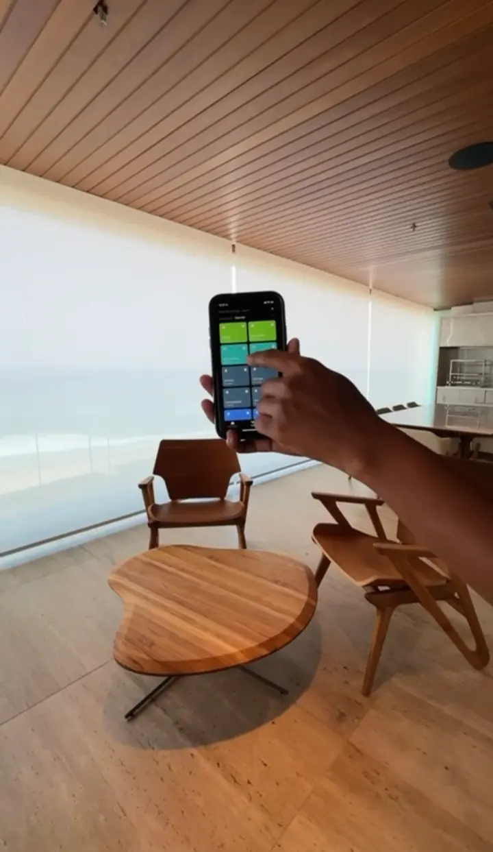 Mão segurando o celular com o app de cenas diante de persianas rolô translúcidas, com o mar ao fundo, em sala com forro de madeira.