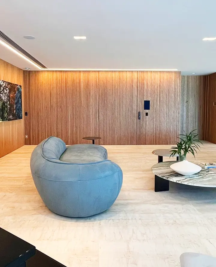 Sala com sofá azul curvo, parede de ripado de madeira, perfil de LED no forro e teclado de controle na parede.